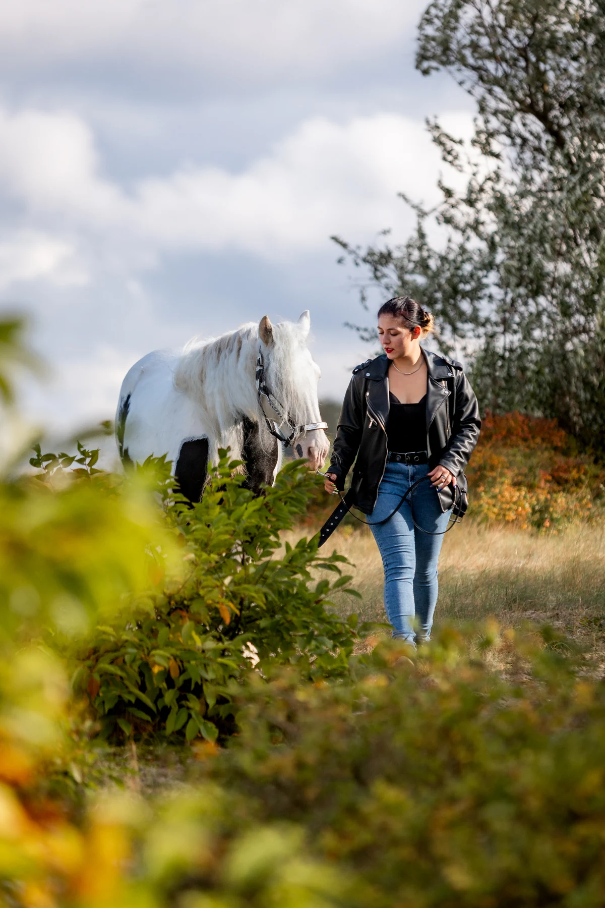 Frau in schwarzer Lederjacke geht mit ihrem gescheckten Pferd durch die herbstliche Natur